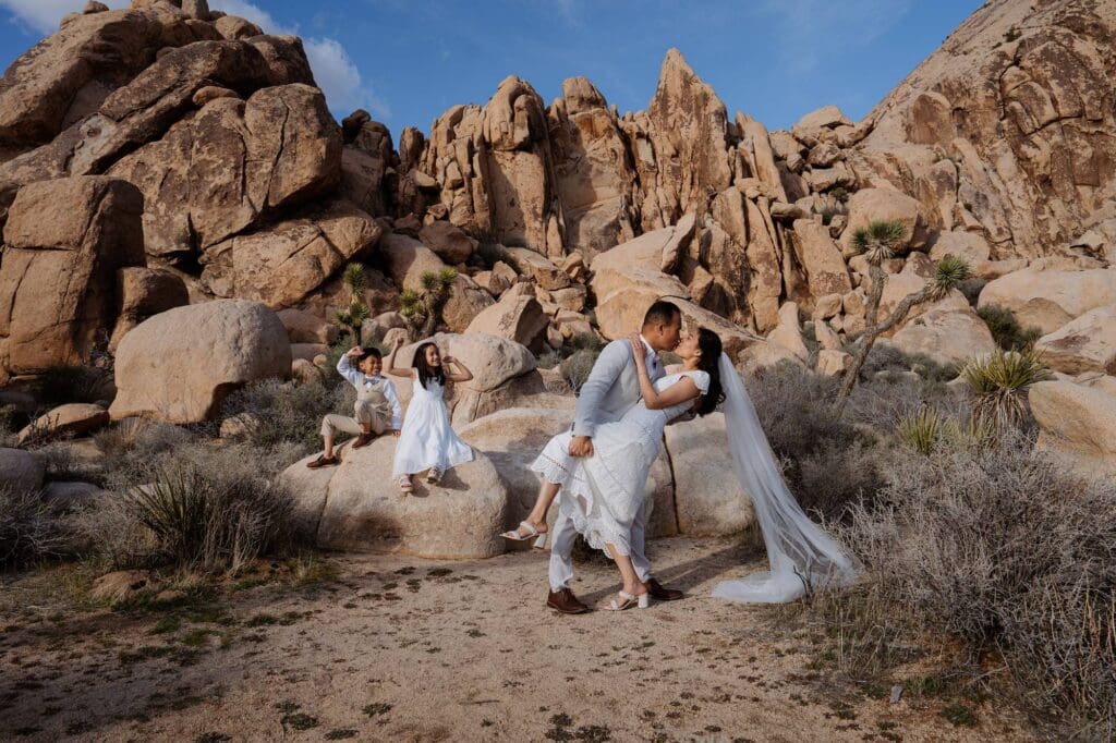 Eloping in Joshua Tree at Hidden Valley – Emerald & Nick Couple exchanging vows while kids watch from a rock ledge