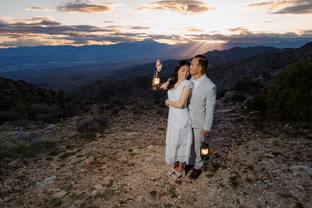 Eloping in Joshua Tree at sunset with lanterns and sweeping desert views Couple eloping in Joshua Tree holding lanterns on a rocky ridge during vibrant sunset at Keys View