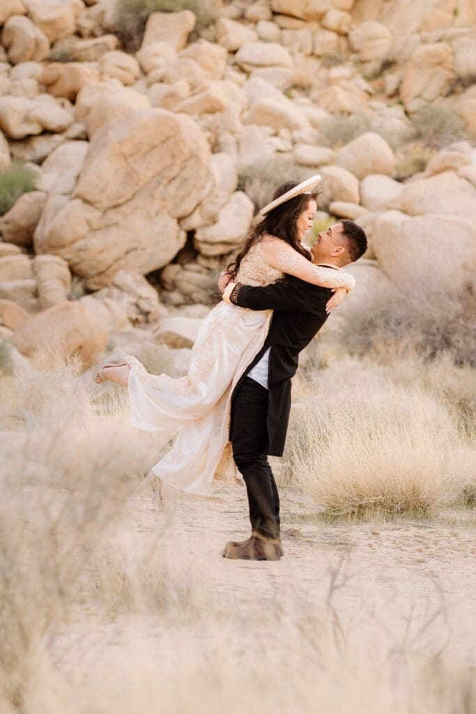Groom lifts bride in Ao Dai during their full-day Joshua Tree elopement surrounded by golden grass and desert boulders