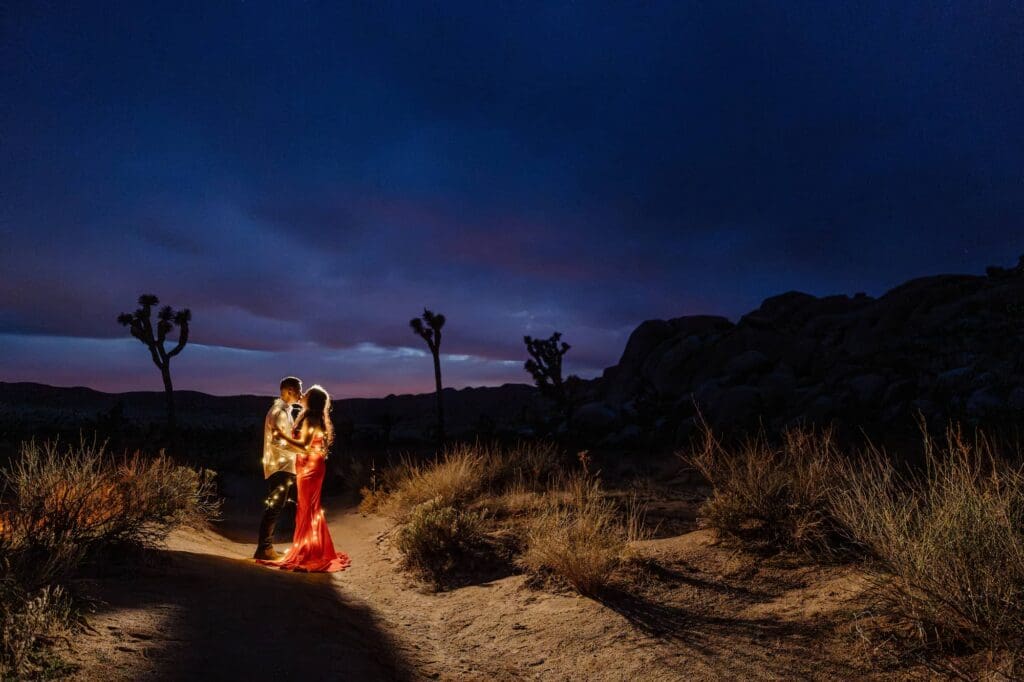 Couple holding guitar under Joshua tree wrapped in fairy lights during blue hour elopement at Cap Rock with evening night sky